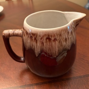 May include: A brown and white ceramic creamer with a drip glaze design. The creamer has a rounded body and a handle.