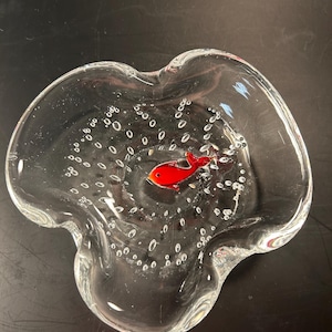May include: A clear glass ashtray with a red fish design in the center. The ashtray has a wavy, organic shape and is decorated with small air bubbles throughout the glass.