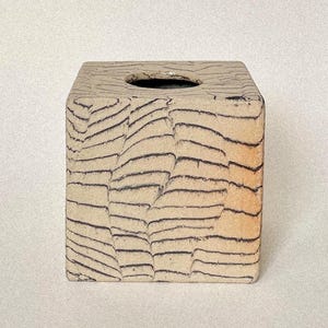 May include: A square-shaped ceramic tissue box cover featuring a textured, light beige surface with dark, hand-drawn horizontal lines that mimic a wood grain pattern. The top surface includes a centred, oval-shaped opening for dispensing tissues.