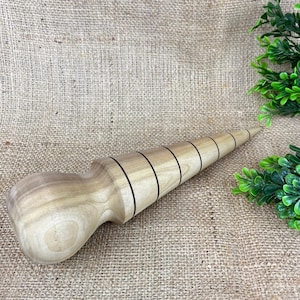 May include: A light-colored wooden massage tool with a rounded handle and a conical, ridged head. The ridges are marked with thin, dark lines. The tool is designed for massage and is made of natural wood.