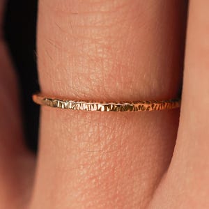 May include: A delicate gold ring with a textured surface. The ring is thin and appears to be handmade. The ring is a warm gold color and is worn on a finger.