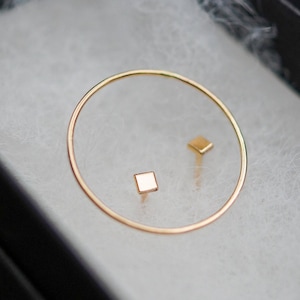 Puede incluir: Un conjunto de dos pendientes de oro, uno es un cuadrado pequeño y el otro es un círculo delgado. Los pendientes se muestran en una caja negra con forro blanco.