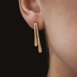 May include: A pair of gold-toned earrings with a minimalist design. The earrings are made of two thin, flat pieces of metal that are connected at the top. The earrings are hanging from the earlobe.