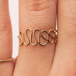 May include: Two gold rings on a hand. The ring on the index finger is a thin, twisted band. The ring on the middle finger is a thicker band with a wavy design.
