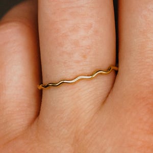 May include: A thin, gold-colored ring featuring a delicate, continuous wavy or rippled design. The band has a smooth, polished metallic finish and maintains a consistent, slender thickness throughout its entire circumference.