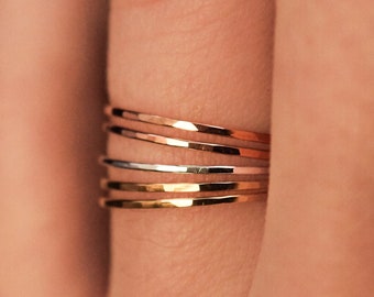 May include: A close-up of a hand wearing a set of four thin gold rings. The rings are stacked on top of each other and are made of different metals, including gold, silver, and rose gold.