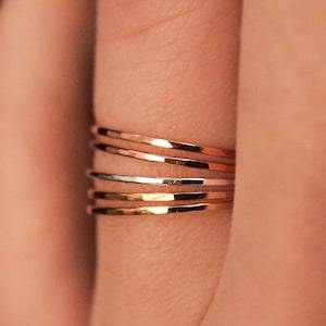 Puede incluir: Primer plano de una mano con un conjunto de cuatro anillos finos de oro. Los anillos están apilados uno encima del otro y están hechos de diferentes metales, incluyendo oro, plata y oro rosa.