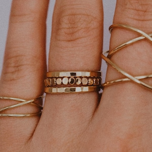 Puede incluir: Una mano con tres anillos de oro. El anillo en el dedo medio es una pila de tres bandas delgadas con una fila de pequeños círculos dorados. El anillo en el dedo índice es una banda gruesa de oro retorcido.