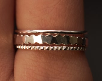 May include: Three silver rings stacked on a finger. The top ring has a scalloped edge, the middle ring has a textured surface, and the bottom ring has a beaded edge.
