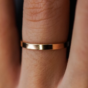 May include: A close-up shot of a gold-colored ring. The ring is a simple band with a flat surface. The ring is on a finger, and the skin tone is light. The ring reflects light, creating a shiny appearance.