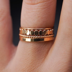 Puede incluir: Un conjunto de tres anillos de oro apilados en un dedo. El anillo superior tiene una fila de pequeñas cuentas redondas de oro texturizado. El anillo del medio es una banda delgada de oro trenzada. El anillo inferior es una banda plana de oro liso.