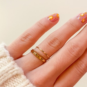 May include: A close-up of a hand wearing three gold rings on the ring finger. The rings are stacked on top of each other and are different widths. The nails are painted with a light pink polish and have small yellow flowers on them.