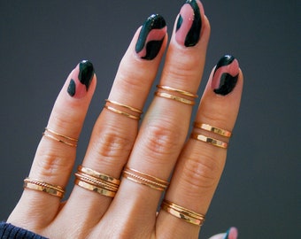 Puede incluir: Una mano con múltiples anillos de oro en cada dedo. Las uñas están pintadas con un diseño de espiral negro y rosa.