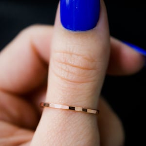 May include: A thin, hammered rose gold band ring worn on a finger with blue nail polish.
