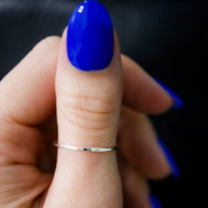 May include: A hand wearing a thin silver ring on the thumb finger. The fingernails are painted a bright blue colour.