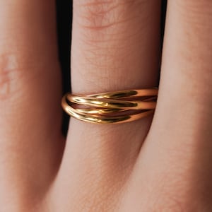 Könnte beinhalten: Nahaufnahme eines goldfarbenen Rings mit einem einzigartigen, ineinandergreifenden Design. Der Ring scheint aus mehreren Bändern zu bestehen, die miteinander verwoben sind. Der Ring befindet sich an einem Finger.