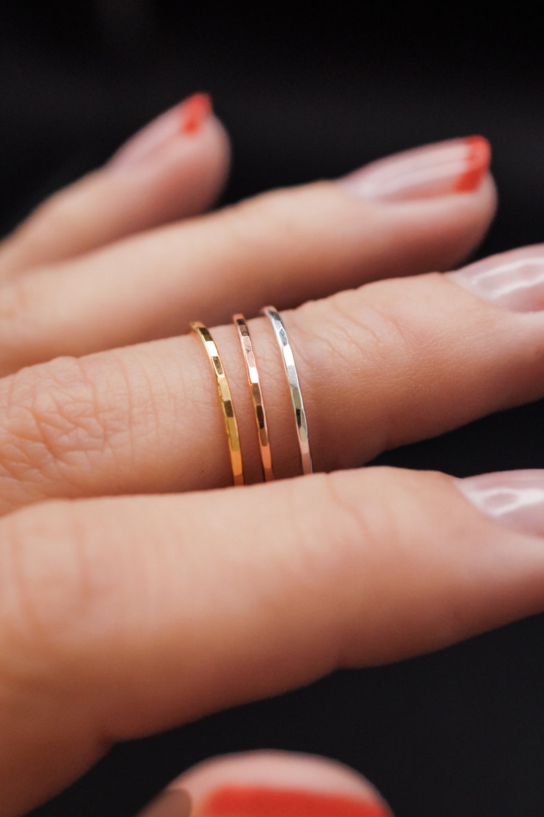 K&ouml;nnte beinhalten: Nahaufnahme von vier d&uuml;nnen, geh&auml;mmerten Metallringen in Gold, Ros&eacute;gold und Silber. Die Ringe sind auf einem Finger vor dunklem Hintergrund gestapelt. Die Ringe sind zart und minimalistisch.