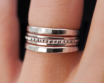 May include: A set of three silver stacking rings. The rings are all different widths and have different textures. The top ring has a hammered finish, the middle ring has a beaded texture, and the bottom ring is smooth.