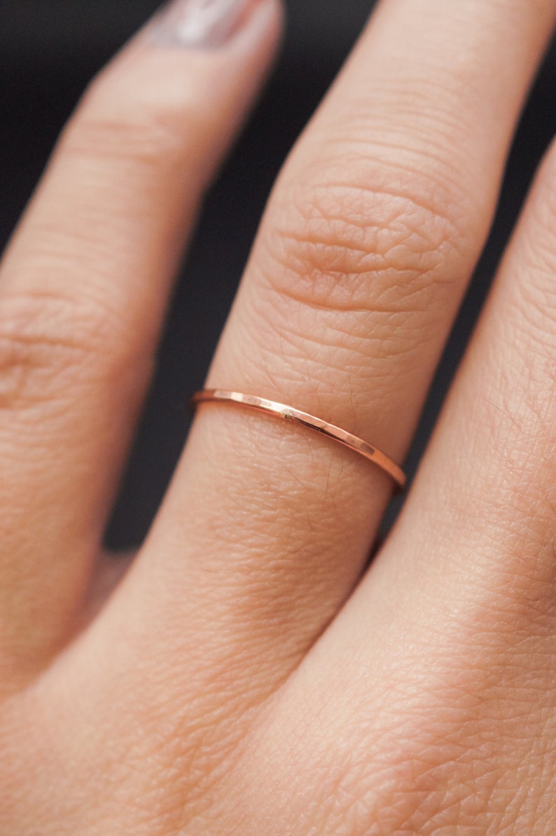 K&ouml;nnte beinhalten: Nahaufnahme eines zarten, d&uuml;nnen ros&eacute;goldenen Rings an einem Finger. Der Ring hat ein schlichtes, minimalistisches Design mit leicht strukturierter Oberfl&auml;che. Der Ring ist im Bandstil gehalten, perfekt zum Stapeln oder einzeln Tragen. Die Farbe des Rings ist ein warmer, metallischer Farbton.