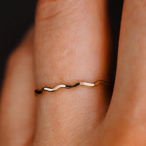 May include: A thin, polished gold ring featuring a continuous, gentle wavy or serpentine design. The band has a smooth, reflective metallic surface that catches the light, creating subtle highlights along the curved, undulating edges of the metal.