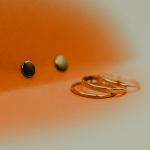 Puede incluir: Tres anillos de oro apilados uno encima del otro sobre una superficie naranja. Dos pequeños pendientes redondos negros están en el fondo.