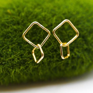 May include: A pair of gold-colored earrings. Each earring features a square shape connected to a smaller, dangling diamond shape. The earrings are set against a vibrant green background.