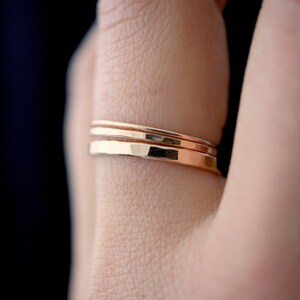 May include: Three thin gold rings stacked on a finger. The rings are smooth and have a slightly hammered texture.