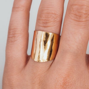 May include: A wide gold band ring with a hammered texture. The ring is worn on a finger.