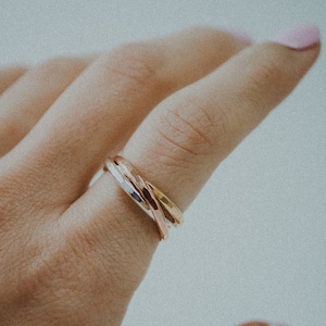 Pode incluir: Um close-up de uma m&atilde;o usando um anel de tr&ecirc;s bandas. O anel &eacute; feito de tr&ecirc;s bandas de metais diferentes: prata, ouro e ouro rosa. As bandas s&atilde;o entrela&ccedil;adas e t&ecirc;m uma textura martelada.