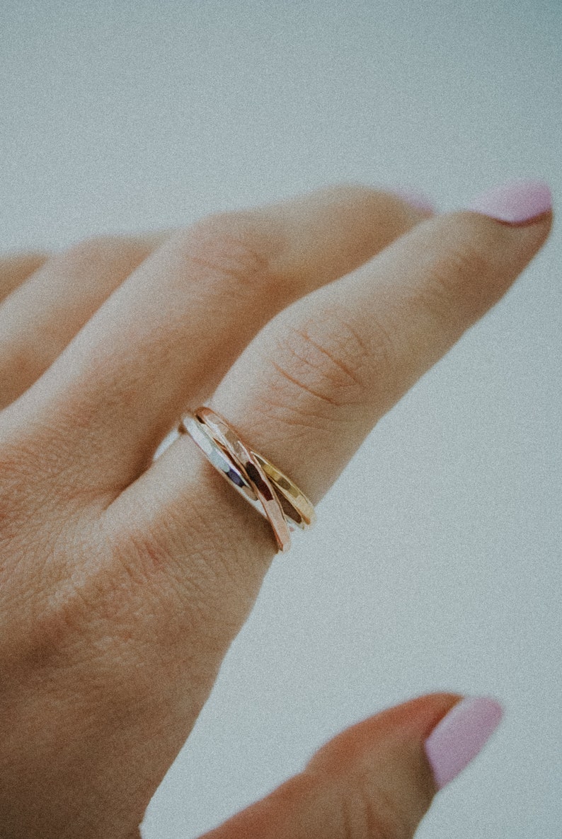 Pode incluir: Um close-up de uma m&atilde;o usando um anel de tr&ecirc;s bandas. O anel &eacute; feito de tr&ecirc;s bandas de metais diferentes: prata, ouro e ouro rosa. As bandas s&atilde;o entrela&ccedil;adas e t&ecirc;m uma textura martelada.