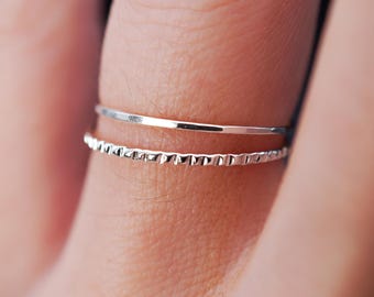 May include: Two silver rings on a finger. The top ring is a simple band, while the bottom ring has a textured design.