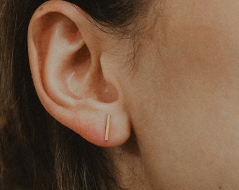 以下が含まれることがあります： 小さな金の棒状のイヤリングをした人の耳のクローズアップ。