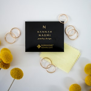 May include: A black jewelry box with gold lettering that reads "HANNAH NAOMI jewelry design" and "SUNSHINE® POLISHING CLOTH". The box is surrounded by gold rings and yellow flowers.