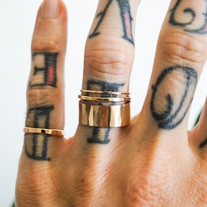 Può includere: Una mano con tre anelli in metallo martellato dorato sull'anulare. La mano è tatuata con vari disegni.