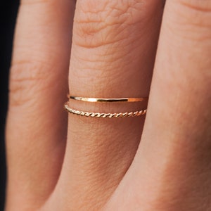 May include: Two gold rings on a finger. The top ring is a thin, flat band. The bottom ring is a twisted band.