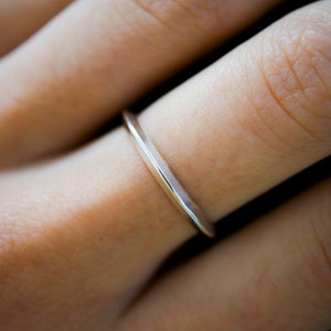 May include: A minimalist silver ring with a rectangular band is worn on a finger. The ring has a sleek, modern design, reflecting light. The background is blurred, focusing attention on the jewellery.