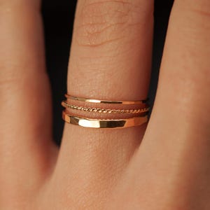 May include: A set of three gold stacking rings. The rings are thin and delicate, with a simple design. The top ring is a thin band, the middle ring is a twisted rope design, and the bottom ring is a flat band.