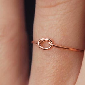 以下が含まれることがあります： シンプルなローズゴールドの結び目のリング。リングは細いワイヤーで作られており、繊細な結び目のデザインが施されています。