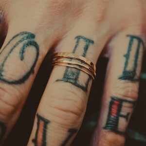 May include: A close-up of a hand wearing three thin gold rings on the ring finger. The hand is tattooed with black ink, featuring the letters "V", "II", and "II" on the fingers.