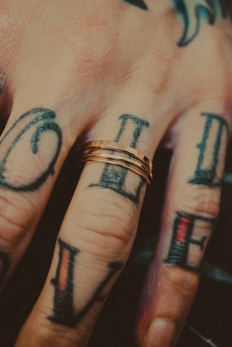 May include: A close-up of a hand wearing three thin gold rings on the ring finger. The hand is tattooed with black ink, featuring the letters "V", "II", and "II" on the fingers.