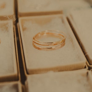 May include: Three gold rings stacked on top of each other in a small, white box. The rings are hammered and have a textured surface.
