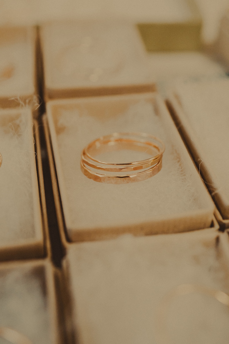 May include: Three gold rings stacked on top of each other in a small, white box. The rings are hammered and have a textured surface.