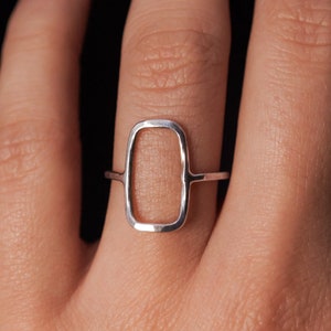 Könnte beinhalten: Ein silberner Ring mit einem rechteckigen, offenen Design. Der Ring wird an einem Finger getragen.