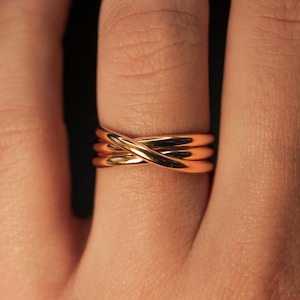 May include: A gold-toned ring with a criss-cross design. The ring is worn on a finger.