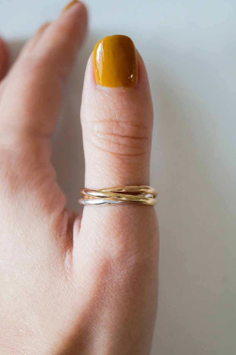 Pode incluir: Um anel de tr&ecirc;s bandas em ouro, prata e ouro rosa em um dedo com esmalte de unha amarelo.