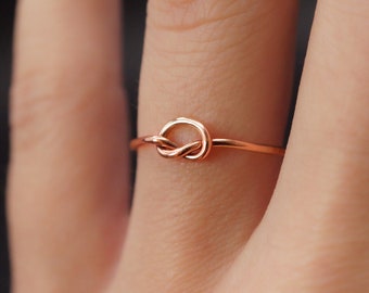 Op de afbeelding: Een delicate roségouden ring met een knoopontwerp. De ring heeft een dunne band en een knoop als middelpunt. De ring wordt getoond op de vinger van een persoon. De ring is gemaakt van roségoud.
