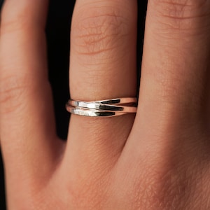 Könnte beinhalten: Nahaufnahme einer Hand mit einem silbernen Dreibandring. Der Ring besteht aus dünnen Bändern, die leicht verdreht und gehämmert sind.