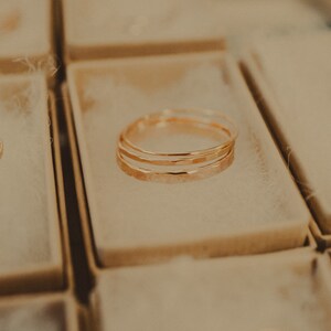 May include: Three gold rings stacked inside a small, beige box. The rings are hammered and have a slightly irregular shape.