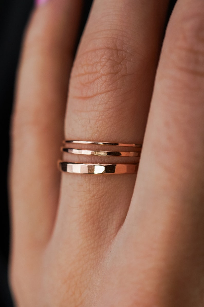 May include: Three gold-toned stacking rings on a finger. The rings are thin and have a hammered texture.