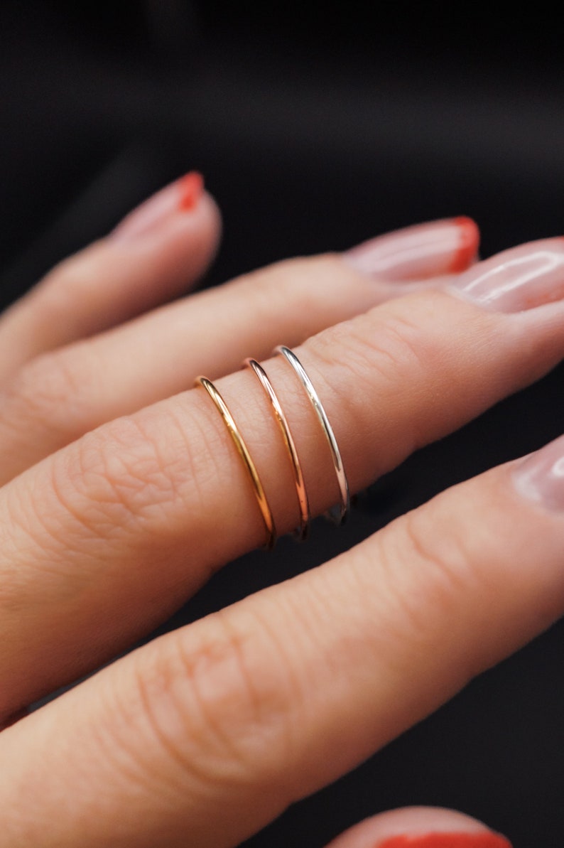 K&ouml;nnte beinhalten: Nahaufnahme von drei d&uuml;nnen Ringen in Gold, Ros&eacute;gold und Silber. Die Ringe werden an einem Finger vor dunklem Hintergrund getragen. Die Ringe sind zart und minimalistisch gestaltet.